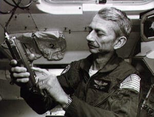 An astronaut in a flight suit with a U.S. flag patch examines a portable ham radio transceiver inside a spacecraft, surrounded by control panels and storage bags.