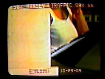 Grainy surveillance photo shows a woman in a white tank top at a counter or partition. Text appears at the top and bottom of the screen, with a date stamp 10-23-05 and 'Munsey Traffic Car 66'.