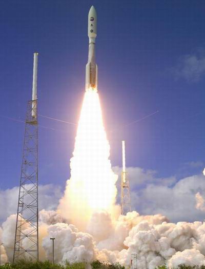 A rocket launches into a clear blue sky, emitting bright flames and smoke from its engines as it ascends, with two tall metal towers on either side and clouds in the background.