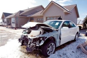 A white car with severe front-end damage is parked on a snowy patch in front of a beige suburban house with a two-car garage. The hood is crumpled and several parts are missing.