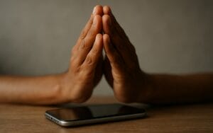 Two hands pressed together in a prayer gesture are positioned above a smartphone lying on a wooden surface, with a neutral, blurred background.