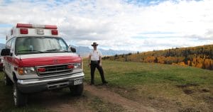 A man in a hat and white EMS shirt stands near a red and white ambulance parked on a grassy hill, with autumn trees and mountains visible in the background under a partly cloudy sky.