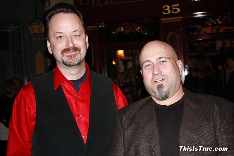 Randy Cassingham and Chris White side by side, smiling at the camera. Randy wears a red shirt and black vest, Chris has a shaved head and a brown jacket. People and festive decor are visible in the background.
