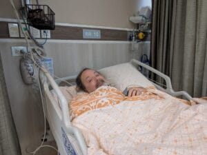 A man with a beard lies in a hospital bed, covered by a patterned blanket. He smiles slightly, surrounded by medical equipment in a hospital room marked “RR 10.”.