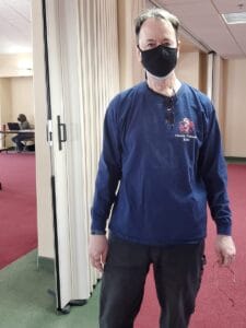 A man wearing a black face mask, dark blue long-sleeve shirt reading "Ouray County EMS", and dark pants stands indoors on a red and green carpet. He is holding glasses and looking at the camera. Another person sits at a table with a computer in the background.