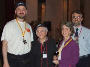 Four adults, all wearing lanyards and access badges, stand together indoors. The woman in the center, Patsy Tombaugh, is older and wears glasses. Randy Cassingham on the left and the central woman wear black Pluto hats. Kit is on Patsy's right, and Rob Staehle is behind her.
