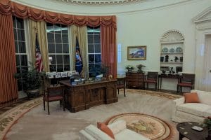 The image shows the Oval Office in the White House with a large wooden desk, U.S. flags, framed photos, a rug with the presidential seal, sofas, a coffee table, and shelves with decorative items along cream-colored walls and tall windows.