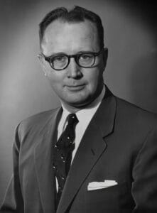 A 1950s black and white professional portrait of a man in a suit and tie wearing round glasses, with neatly combed hair, posing against a plain background. He has a neutral expression and a pocket square in his jacket.