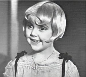 A young girl with short, light-colored bobbed hair and bangs smiles, wearing a light-colored dress with dark straps and a necklace, in a black and white photo.