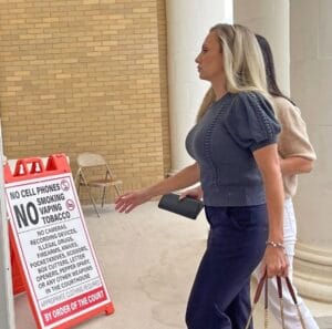 Two women walk by a courthouse sign of banned items like cell phones. The sign stands near brick and columns. One holds a wallet and phone; the other has a brown purse.