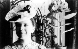 A young woman smiles while wearing a large, lacy hat decorated with a ribbon. She stands indoors, with decorative plants and an ornate screen in the background. The image is in black and white.