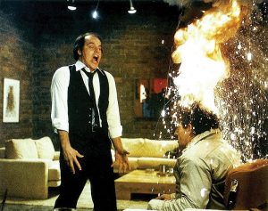 A man in a vest screams in shock as another mans head erupts in flames and sparks in a living room with a sofa and art on the wall in the background from a scene in the movie, Scanners.