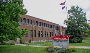 A two-story brick school building with an American flag flying above. In front, a sign reads PS Joyce Kilmer School #69 and advertises a summer camp. Green trees and grass surround the school.