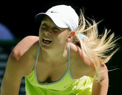 Maria Sharapova playing tennis in a white cap and yellow sports outfit leans forward, appearing focused and intense, with her blonde hair flying back during a match.