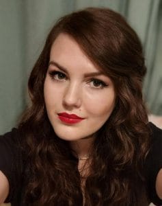 A woman with long, wavy brown hair and red lipstick looks at the camera. She is wearing a black top and has a neutral background behind her.
