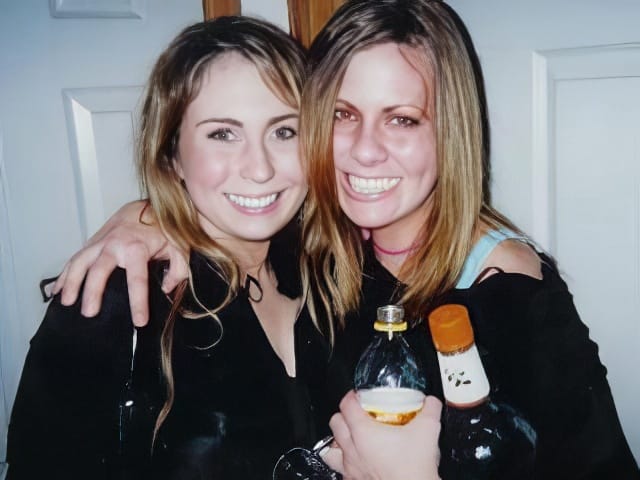 Two young women smiling with their arms around each other. One holds a bottle of juice and a liquor bottle; her skin is flushed, and it appears she has a long scrape on her forehead. They are standing in front of a white door.