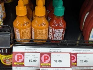 Bottles of “Crying Thaiger” mayo chili and sriracha chili sauces, arranged on a store shelf. Price tags in both Thai and English are displayed below the bottles, indicating promotional discounts.