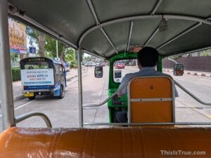 View from the backseat of a tuk-tuk, with the driver visible in front and another tuk-tuk ahead on a street bordered by trees, cars parked alongside, and a sidewalk in the background.