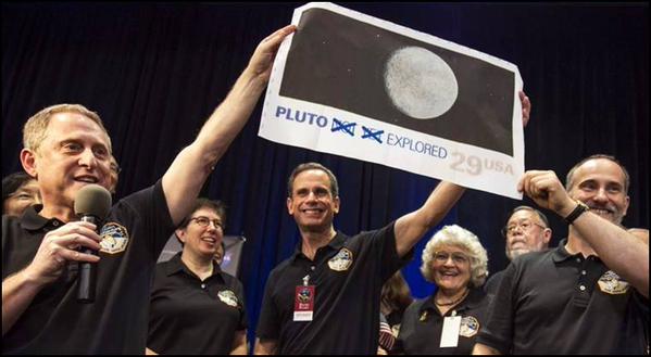 A group of people in black shirts celebrate while holding a banner that reads PLUTO EXPLORED and shows an image of Pluto. One person speaks into a microphone. Everyone is smiling, and the atmosphere is joyful.