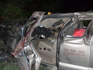 A heavily damaged silver pickup with its driver-side door bent outward and the interior exposed, from a rollover, at night with broken glass and crumpled metal visible.