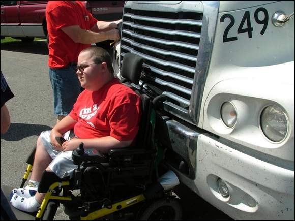 A person in a wheelchair is pinned against the front of a large white truck. Another person stands nearby. The person in the wheelchair appears calm and is wearing a red shirt.