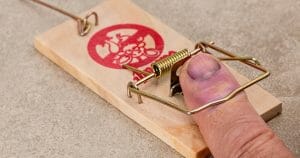 A swollen, bruised finger is caught in a metal mousetrap on a wooden base, lying on a gray surface. The finger appears injured and purple from being trapped.