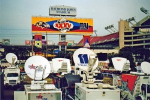 Broadcast vans with satellite dishes are parked outside Raymond James Stadium, with a large Super Bowl XXXV banner featuring the Baltimore Ravens and New York Giants displayed in the background.