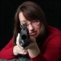 A woman with long brown hair and glasses, wearing a red top, aiming a rifle toward the camera against a dark background.