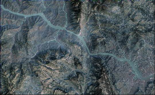 Satellite image showing a winding river cutting through rugged, mountainous terrain with steep ridges and valleys; the landscape appears mostly barren with shades of brown and gray.