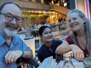 Three people smile together at an outdoor restaurant table in the evening, illuminated by warm lights and surrounded by modern decor. Plates and a glass bottle are on the table. The scene has a friendly and relaxed atmosphere.