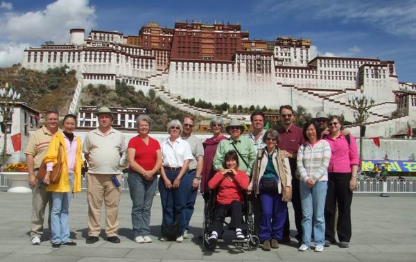 A group of people, including one person in a wheelchair, pose for a photo in front of the Potala Palace in Lhasa, Tibet, on a sunny day with blue skies and scattered clouds.