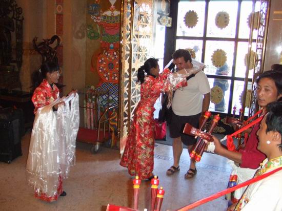 A woman in traditional Chinese dress drapes a white scarf around the neck of a man inside a decorated room, while others in similar attire watch and hold red instruments. Bright light shines through a glass door behind them.