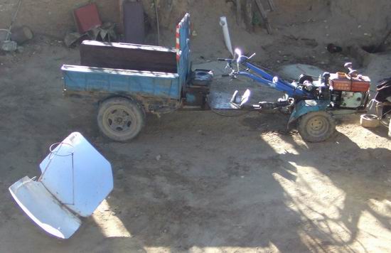 A small, blue, open-bed vehicle with a front resembling a walking tractor is parked on a dirt ground. Nearby, a large, reflective metal panel lies angled on the ground.