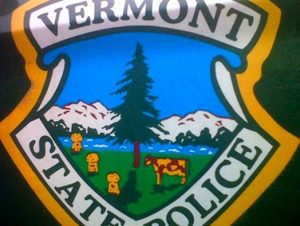 Vermont State Police emblem showing a tree, cows, and gently rolling hills with mountains and a clear blue sky in the background.