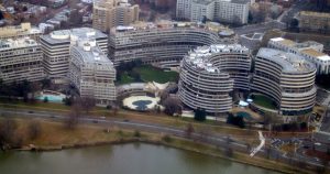 Aerial view of the curved, modernist Watergate complex buildings in Washington, D.C., surrounded by roads, trees, and a river in the foreground.