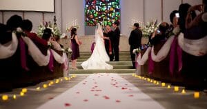 A bride and groom stand at the altar during a wedding ceremony in a church, with guests seated on either side, flower petals on the aisle, and a colorful stained glass window in the background.