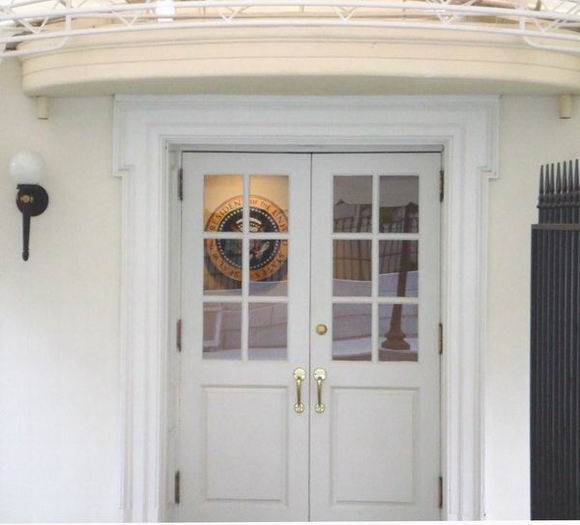 Double white doors with glass panes and brass handles, leading into the White House. The seal of the President of the United States is visible through the glass panes on the wall inside. A wall lamp and black metal fence are seen to the sides.