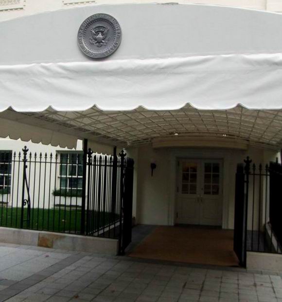 White canopy with the U.S. presidential seal attached covers an entrance to the White House, surrounded by a black iron fence and a green lawn visible through the fence. The double doors appear not to be the main entrance.