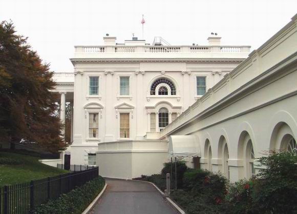 The exterior of the White House, featuring white columns, arched windows, and a pathway lined with greenery leading up to the historic building.