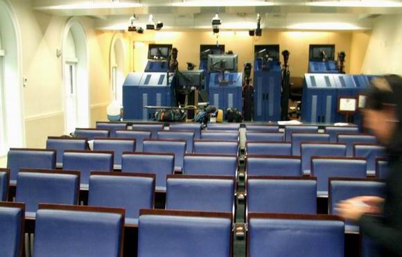 Rows of empty blue chairs face a bank of cameras and equipment in the White House Press Briefing Room. The room is lit by overhead lights, and a person is blurred in the foreground on the right side.
