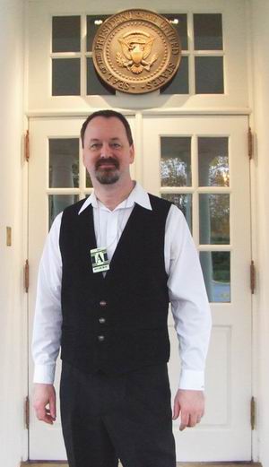 Randy Cassingham with short hair and a goatee, wearing a white shirt and black vest, stands in front of the White House doors with glass panes and a round plaque with the U.S. presidential seal above the doorway.