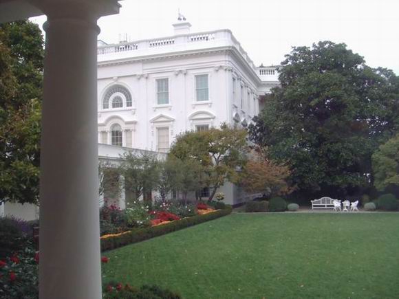 White House exterior with landscaped garden, green lawn, colorful flower beds, trees, and a white bench with chairs on the right side. A large white column is visible in the foreground on the left.