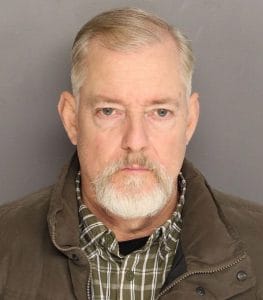 A middle-aged man with short gray hair and a trimmed white beard, wearing a plaid shirt and a brown jacket, poses against a plain gray background.