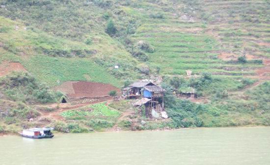 A wooden house stands on a green hillside with terraced fields, next to a river with a boat floating near the shore. The scene is rural and peaceful.