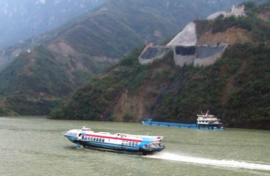 A blue and white high-speed boat travels on a river, with a cargo boat further behind. Green hills and steep slopes with construction sites are visible in the background.