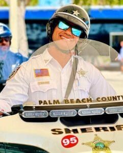A smiling Palm Beach County sheriff’s deputy, wearing a uniform and sunglasses, sits on a motorcycle, while another officer stands in the background.