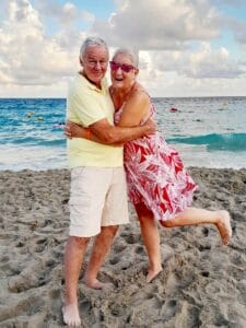 An older gray-haired couple on a beach, standing barefoot in the sand embracing. Paul, in a yellow Polo shirt and beige cargo shorts, is smiling wryly. Joyce, in a red thigh-length dress with a white floral "island" print, appears to be laughing. Behind them is the ocean gently rolling in. Non-threatening clouds obscure most of the blue sky. It's a very nice day at the beach.