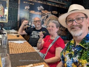 At a restaurant bar. Dan and Kit have sake cups. The place is exotically decorated with a very fancy wall right behind Dan with Asian art. Each of the three have nice bamboo placemats. Kit is wearing a red dress. Randy is smiling, wearing a white Panama hat and tropical motif shirt.