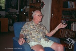 Clarke in his den, seated in a blue upholstered chair, his desk and computer in the background. He is speaking to someone off-camera, gesturing with one hand. A bookshelf next to him includes a set of the Encyclopedia Britannica. (Dan Sokol)