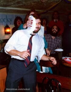 Steve Wozniak, standing at a restaurant. It's obviously a party atmosphere, with a big sign that says "Rocky Racoon" (the rest is cut off). People in the background are looking at him and laughing since directly behind him, a woman is smashing a paper plate filled with whipped cream onto the side of his head. Her hand holding the plate have long, very red nails. Woz is laughing, as are several people in the background. One man is holding a backup plate of whipped cream, if needed.